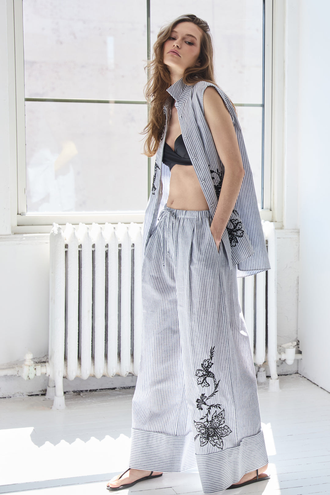 Woman wearing a sleeveless embroidered top and wide-leg pants in a bright room.