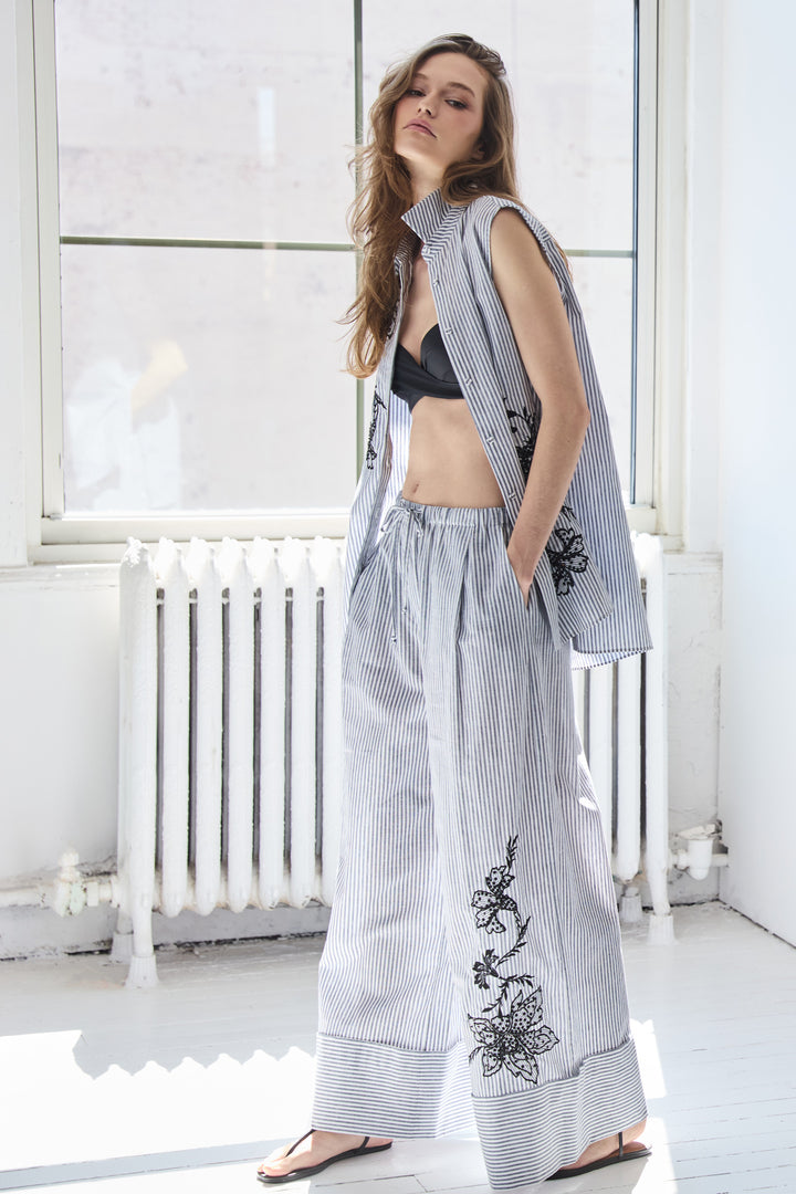 Woman wearing a sleeveless embroidered top and wide-leg pants in a bright room.