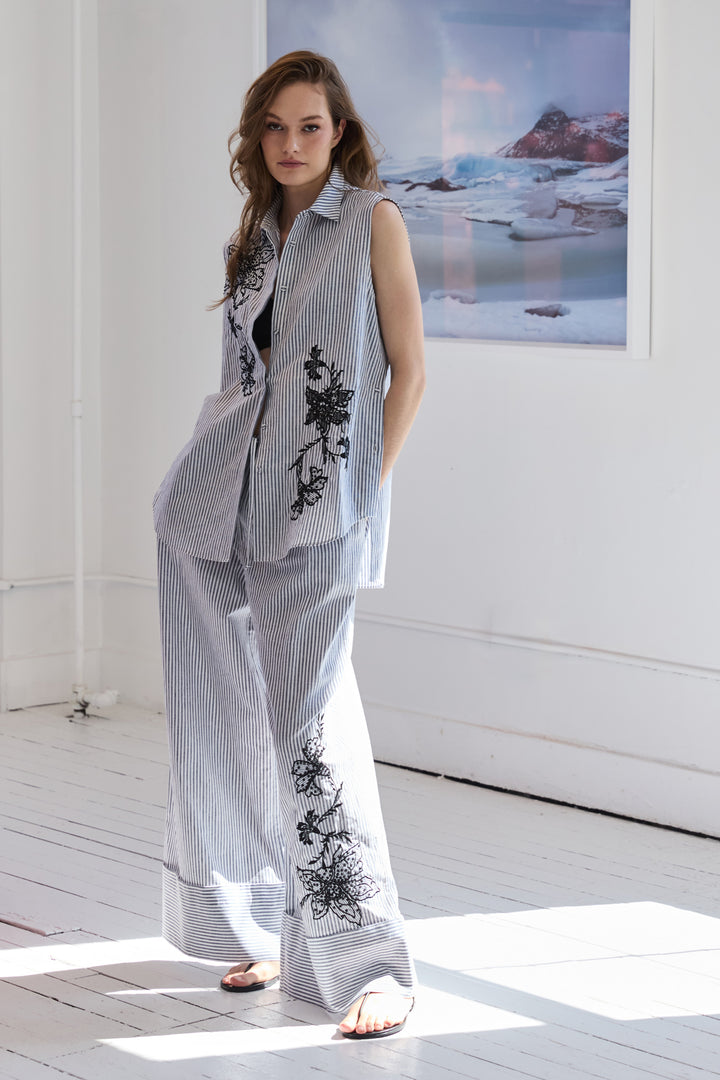 Woman wearing a sleeveless embroidered top and pants in a room with a large window.