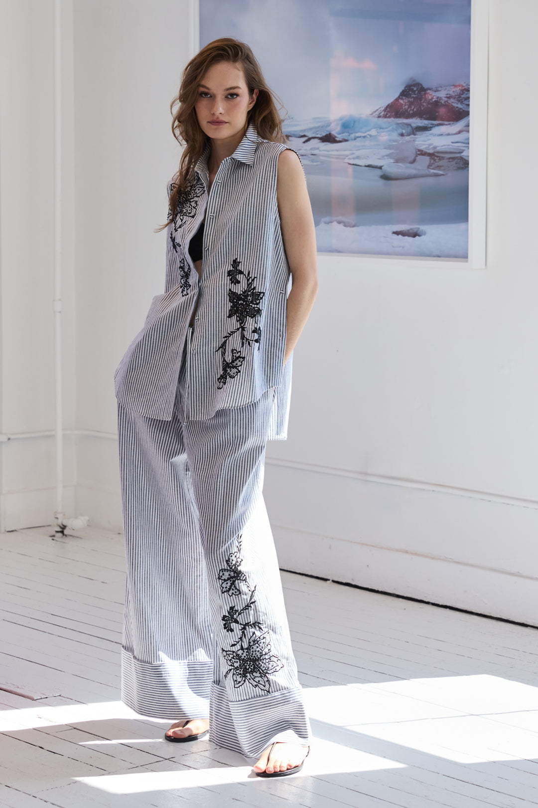 Woman wearing a sleeveless embroidered top and pants in a room with a large window.