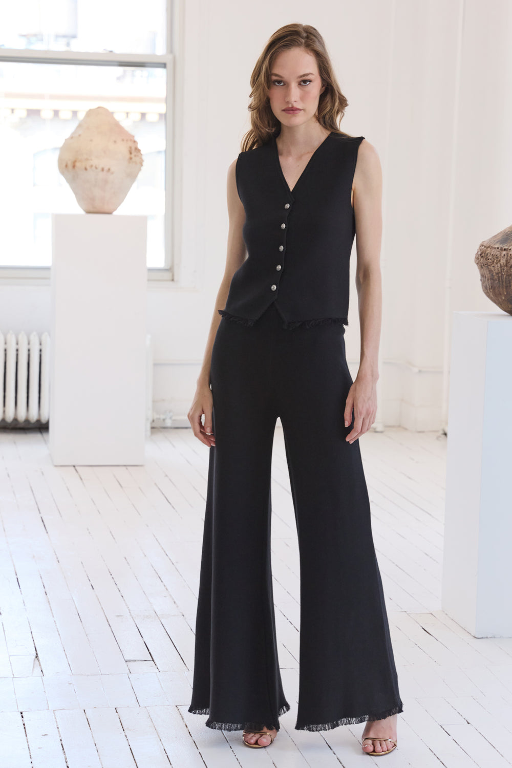 Woman wearing a black sleeveless top and flared pants in a bright room.