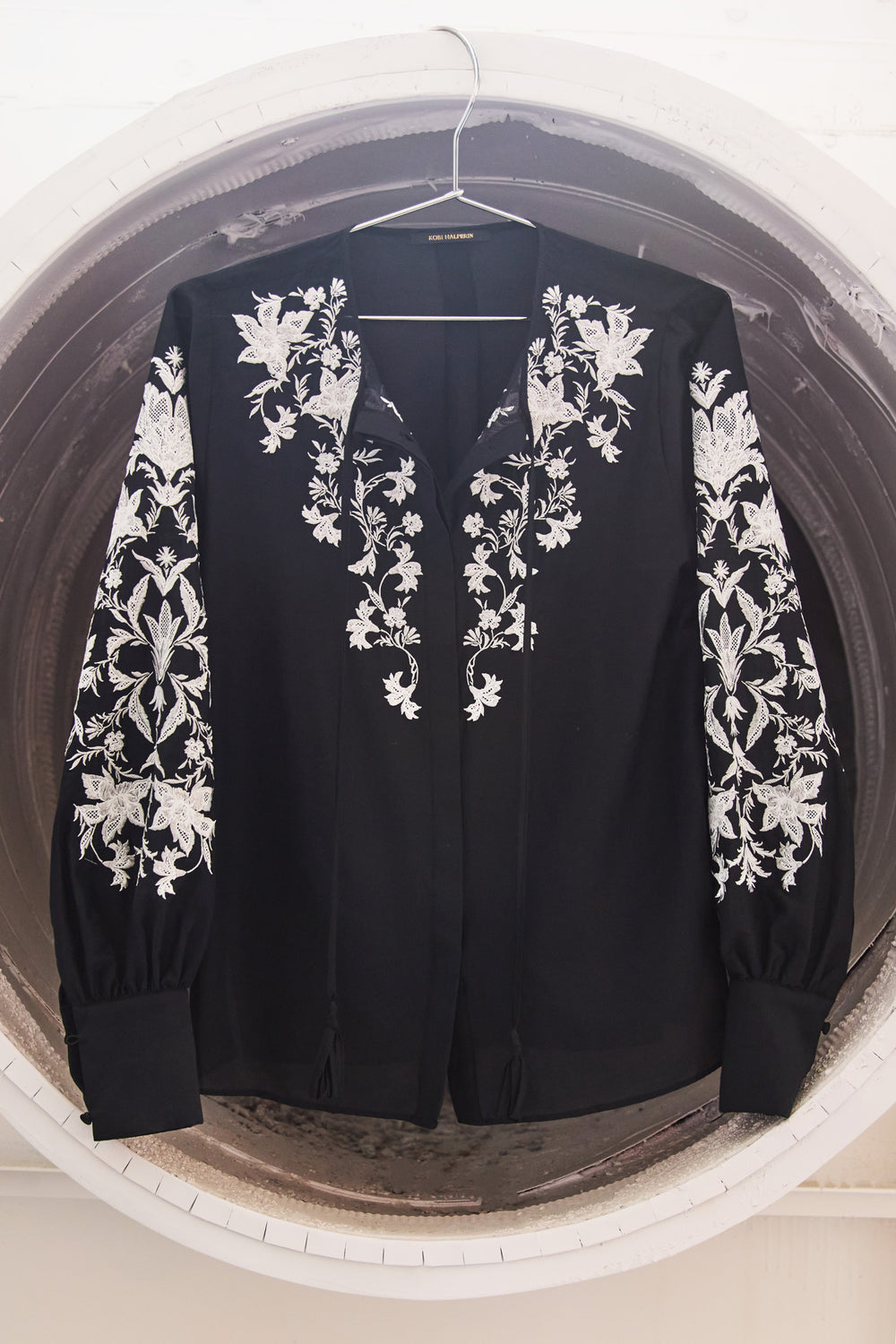 Black jacket with white floral embroidery on a hanger against a neutral background