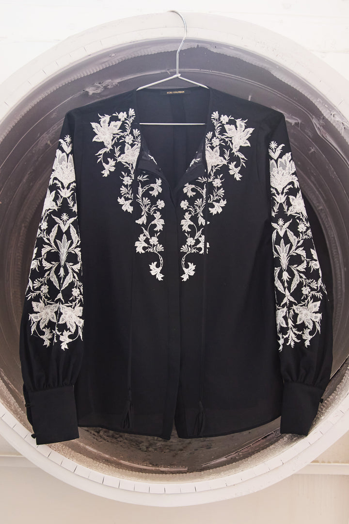 Black jacket with white floral embroidery on a hanger against a neutral background
