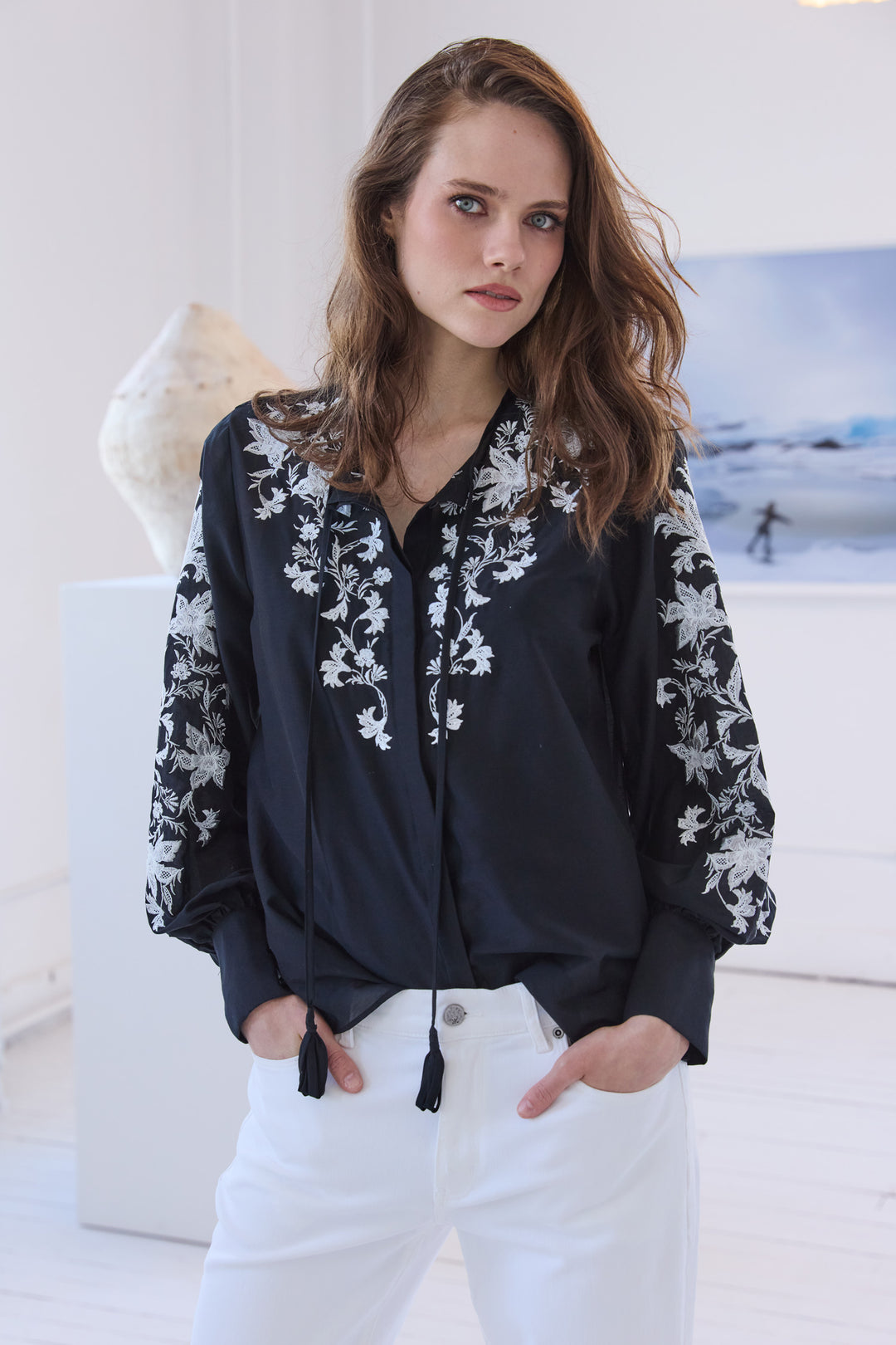 Woman wearing a black blouse with white floral embroidery and white pants in a bright room.