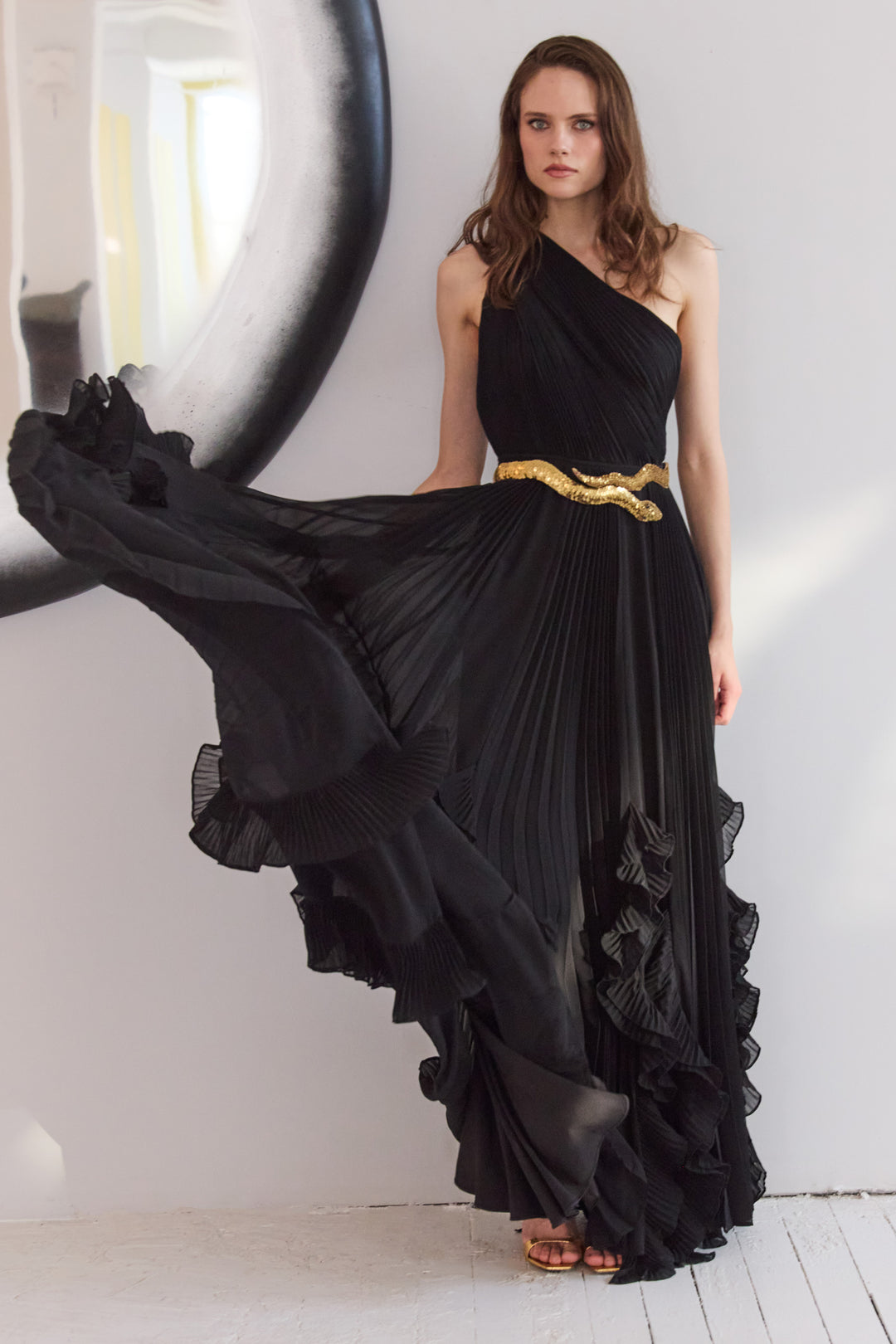 Woman wearing a black one-shoulder dress with ruffled layers and a gold belt, standing against a white wall.