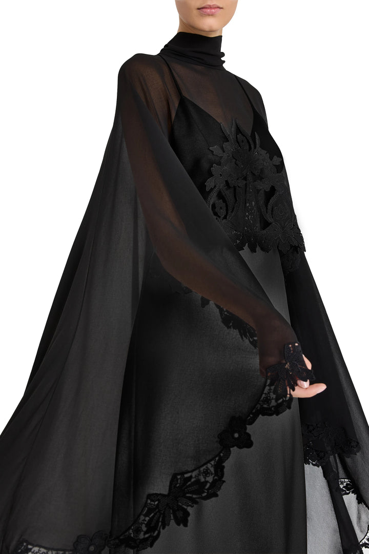 Sheer black, floor-length dress with cape sleeves, lace floral details, and high neckline; front view on model.