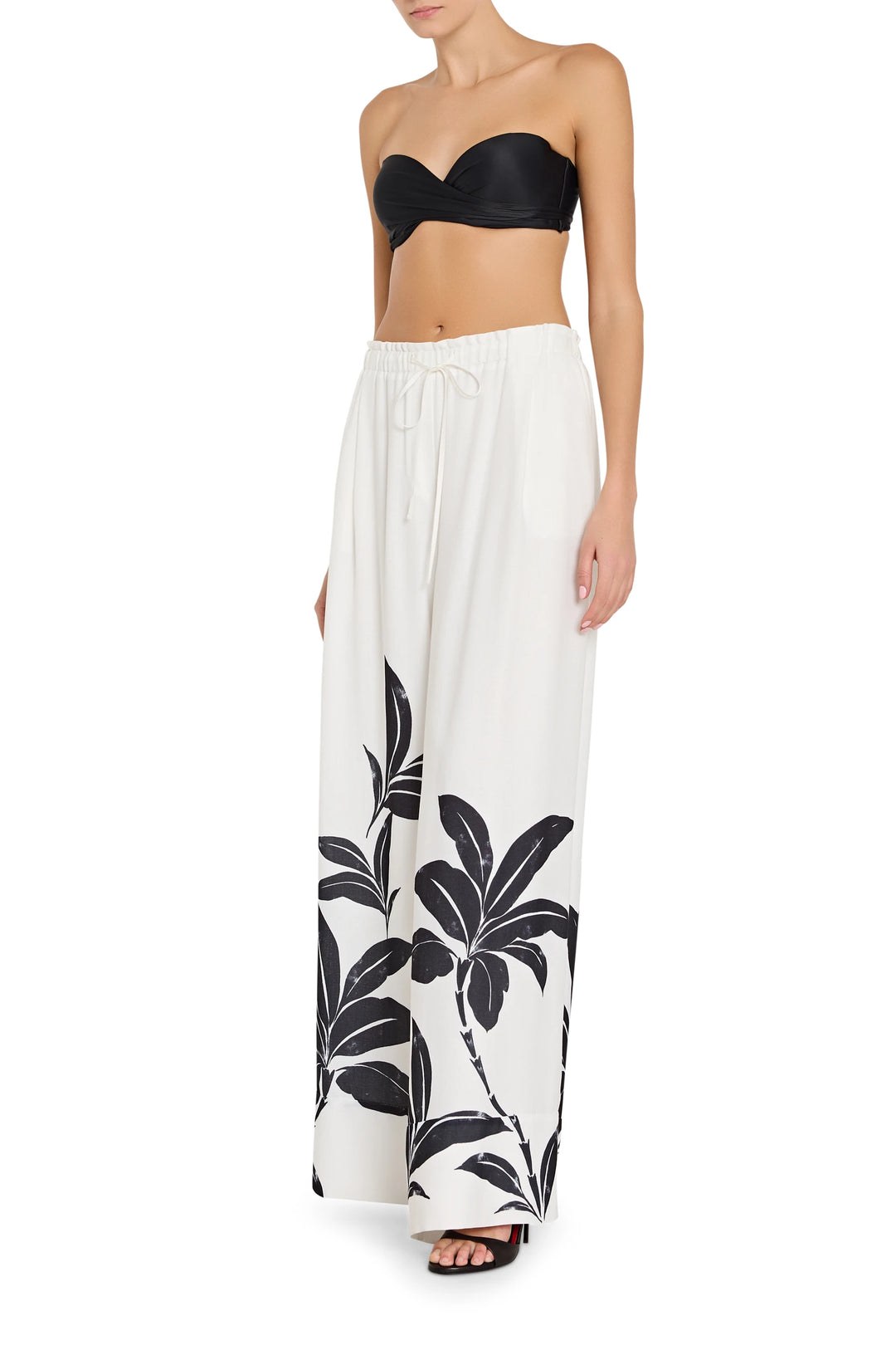 Woman wearing a black strapless top and white pants with black palm leaf patterns on a white background