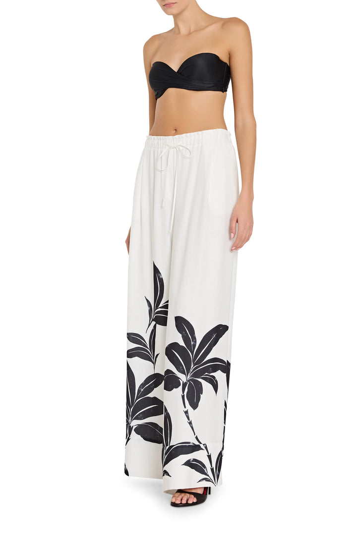 Woman wearing a black strapless top and white pants with black palm leaf patterns on a white background