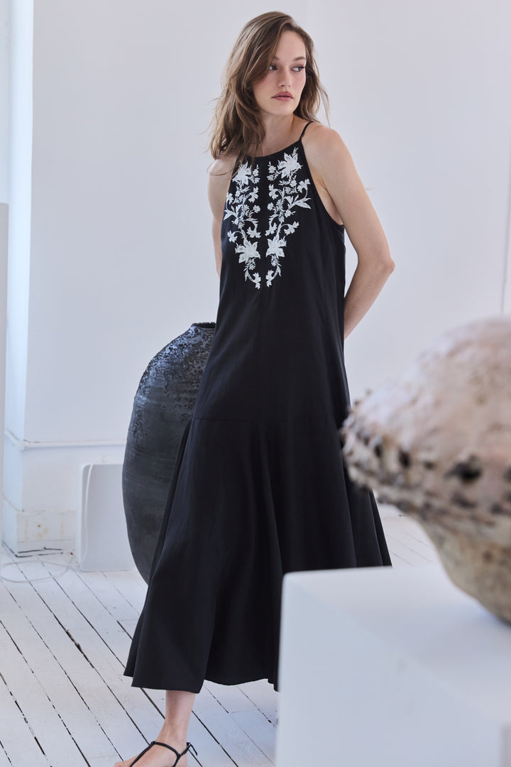 Woman wearing a black sleeveless dress with white floral embroidery in a minimalistic setting.
