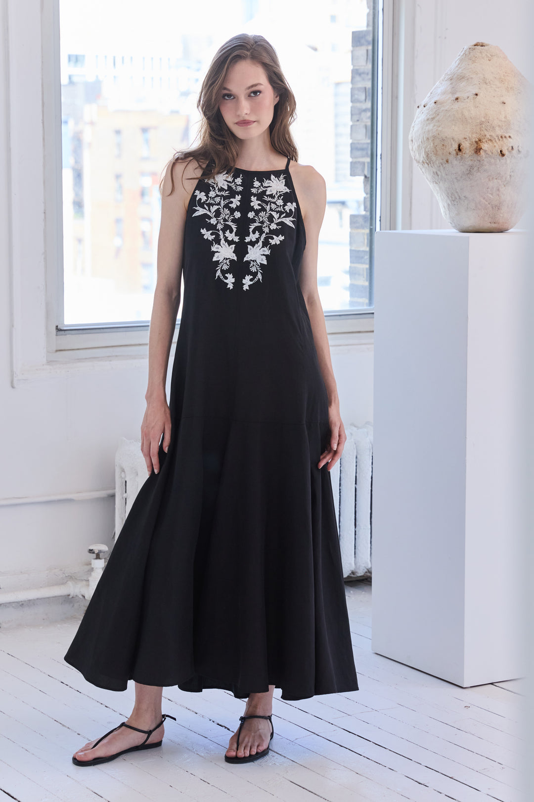 Woman wearing a black sleeveless dress with white floral embroidery in a bright room.