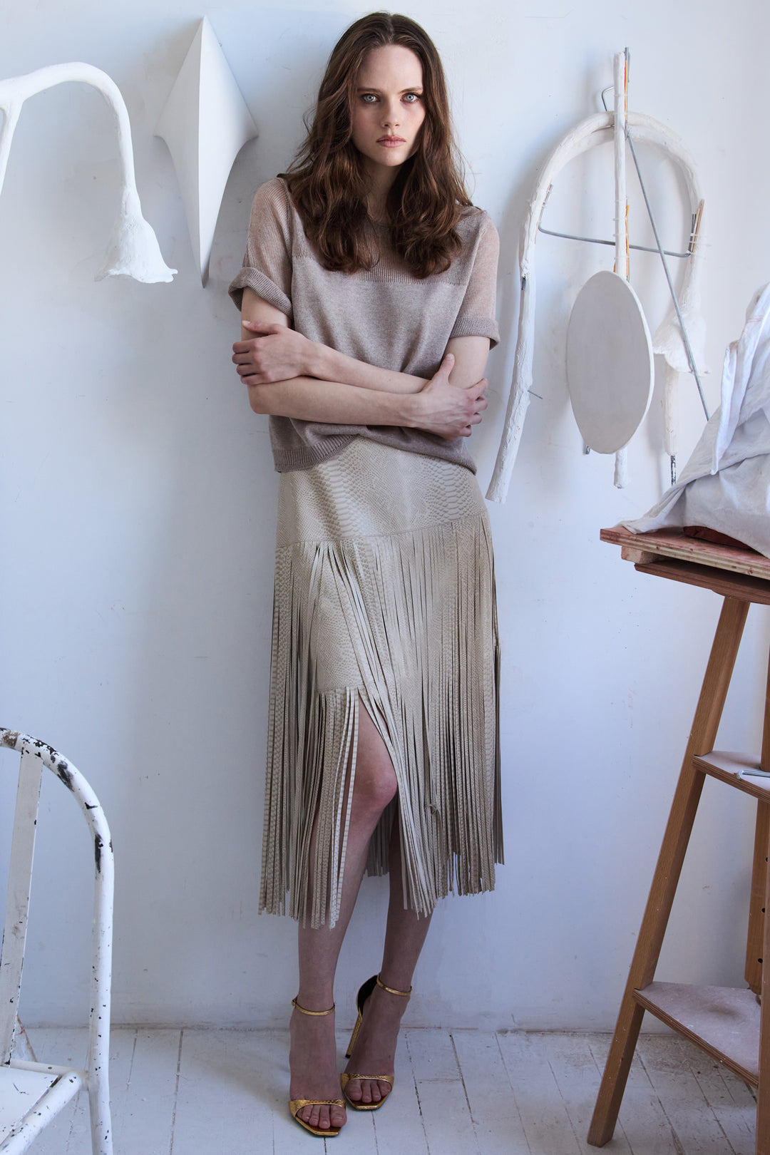 Woman wearing a beige top and fringe skirt standing in a minimalistic room.
