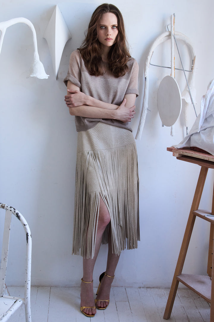 Woman wearing a beige top and fringe skirt standing in a minimalistic room.